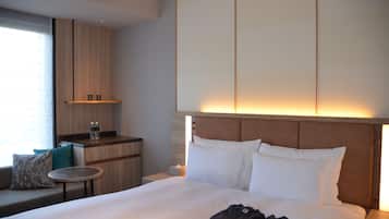 Ein Hotelzimmer mit einem großen Bett, einem kleinen Tisch, einer Couch, einer Minibar und einem Fenster mit Jalousien.
