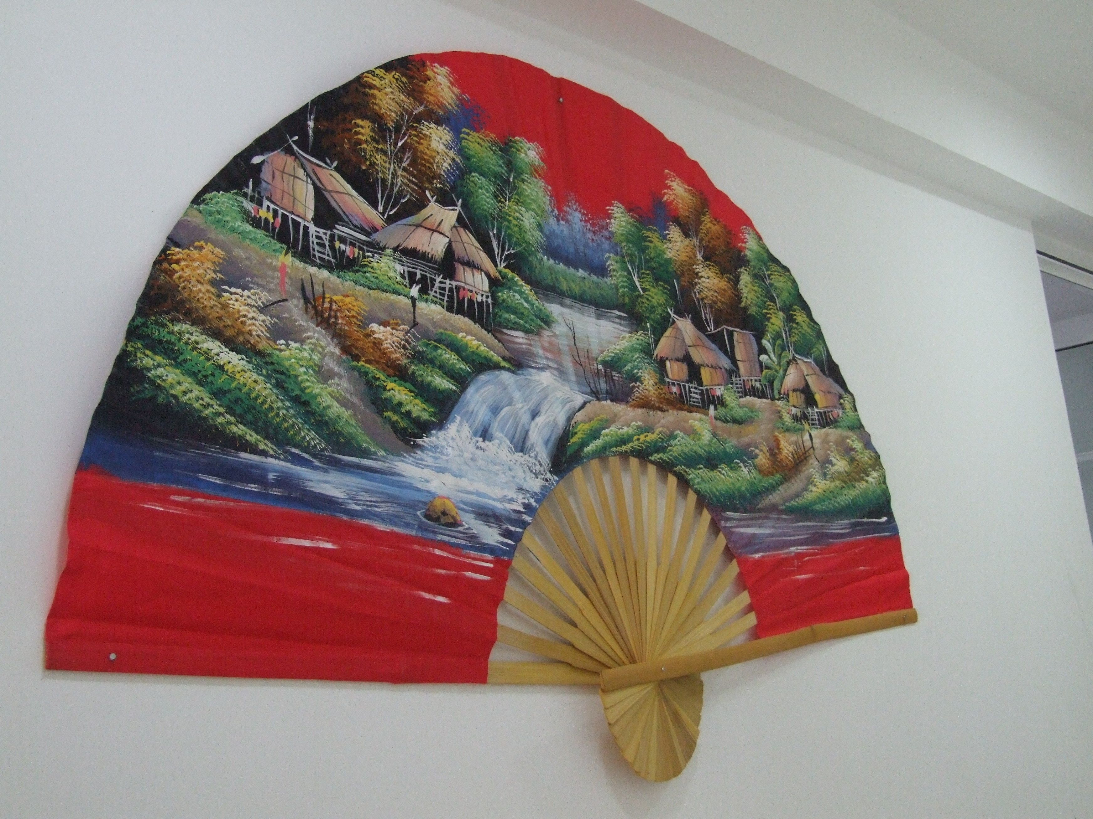 A wall with a painting of a village scene, a fan, and a small object on the floor.