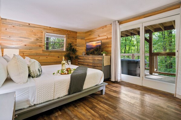 A bedroom with a bed, a dresser, a television, and a large window with a view of trees.