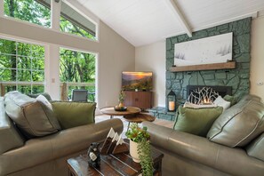 Ein gemütliches Wohnzimmer mit Ledersofas, einem Kamin und einem großen Fenster mit Blick auf einen Wald.
