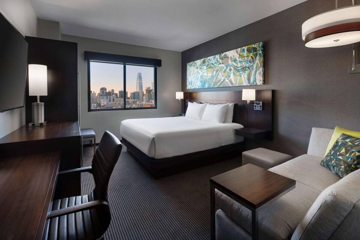 A hotel room with a bed, a desk, a sofa, a large window with a city view, and a painting on the wall.