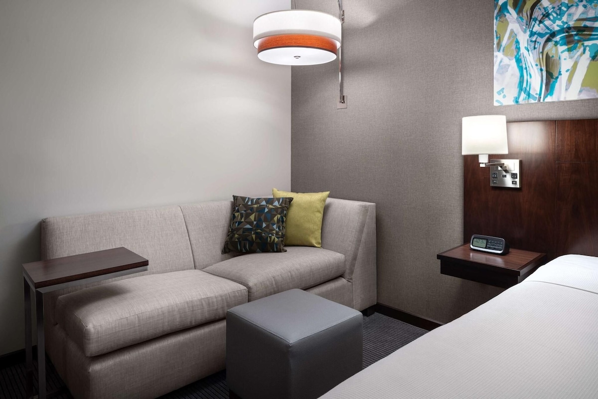 A hotel room with a sofa, a small table, a bedside table with a lamp, and a wall-mounted alarm clock.