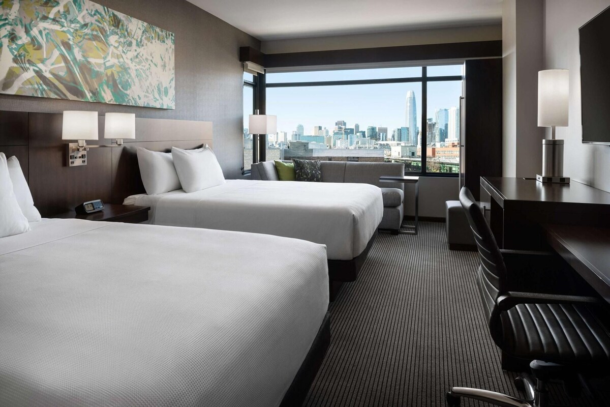 A hotel room with two beds, a desk, a chair, a TV, and a city view.