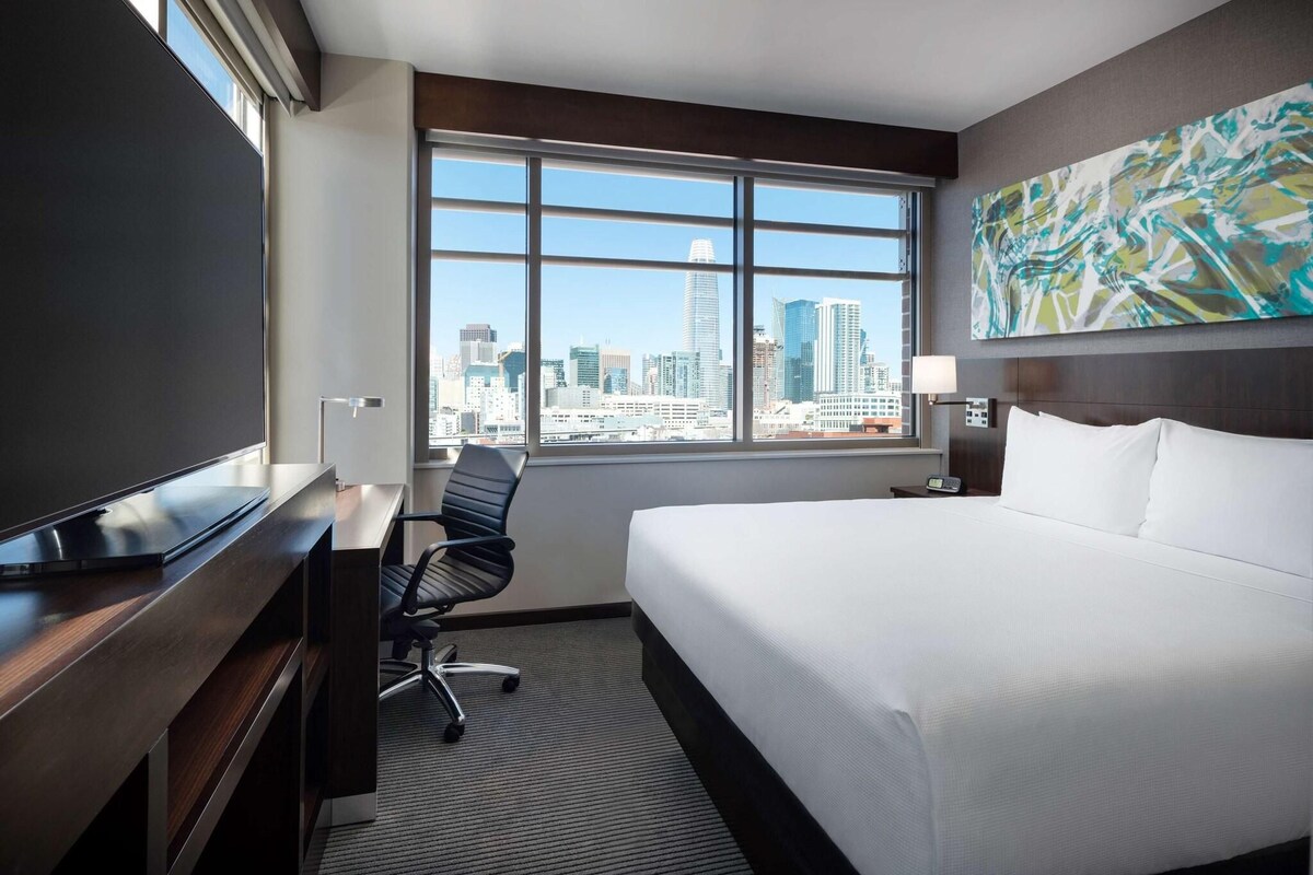 A hotel room with a bed, desk, chair, and a large window offering a city view.