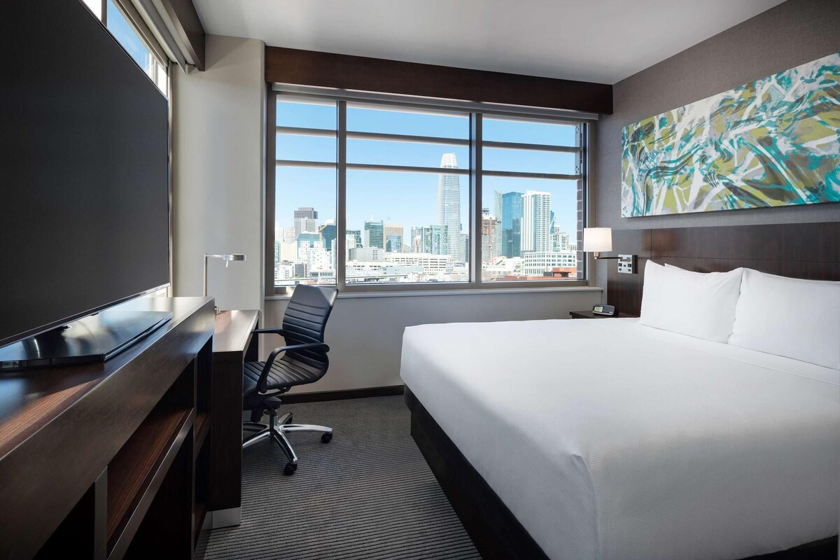 A hotel room with a bed, desk, chair, and a large window with a city view.