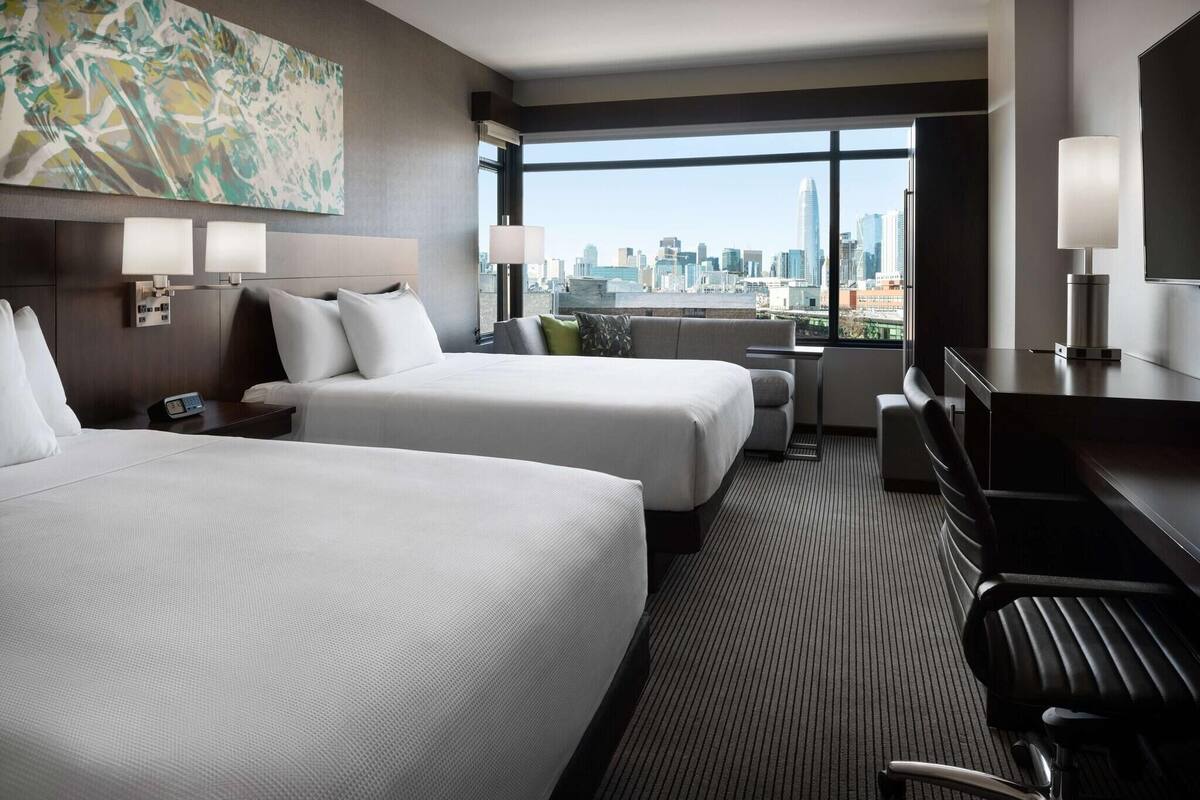A hotel room with two beds, a desk, a chair, a TV, and a city view.