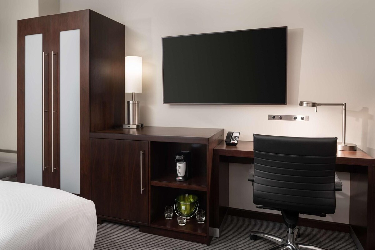 A hotel room with a bed, a desk with a chair, a television, a cabinet with a coffee maker, and a lamp.
