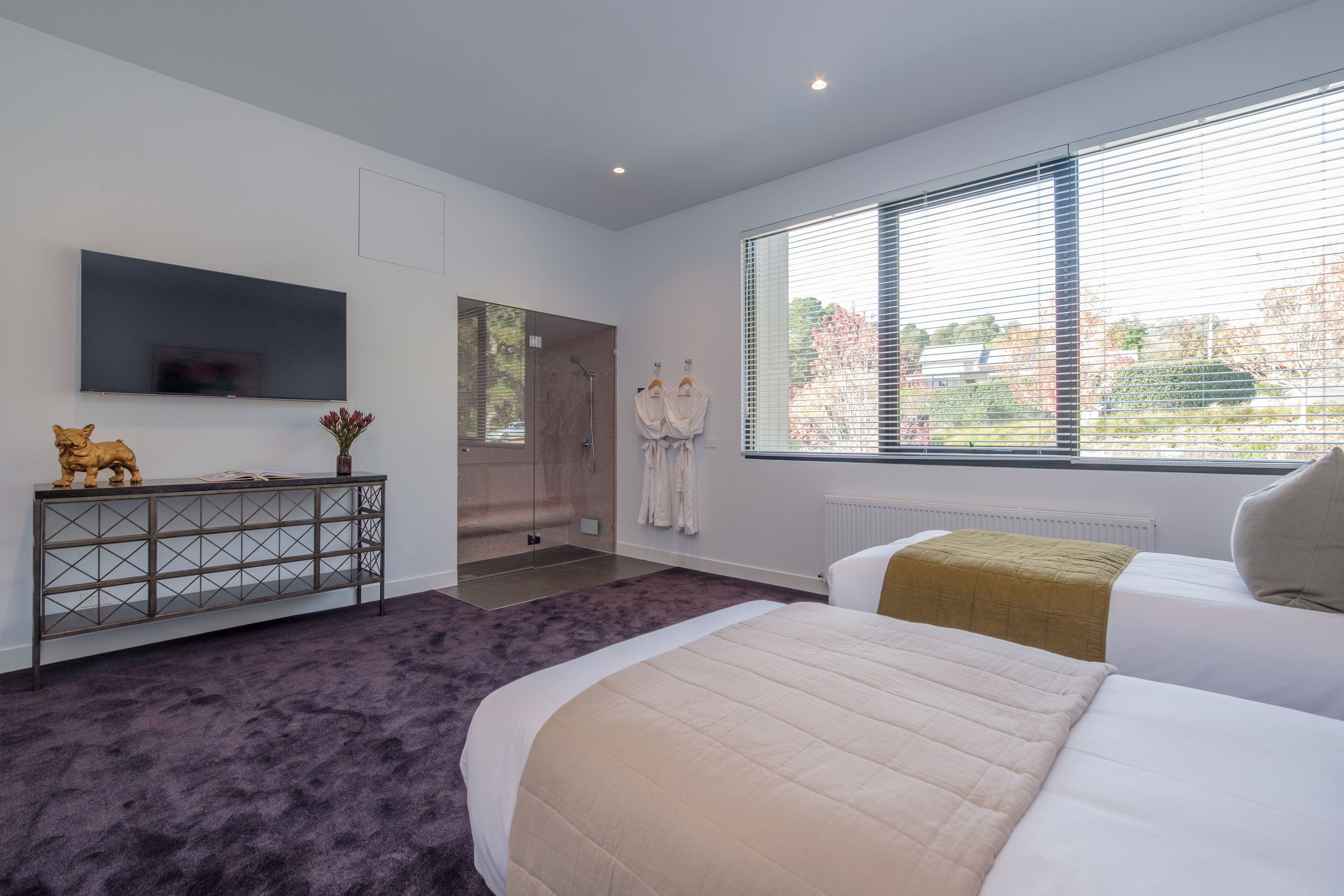 Une chambre moderne avec un grand lit, un téléviseur fixé au mur, une console sur laquelle est posé un objet décoratif, et une fenêtre équipée de stores.