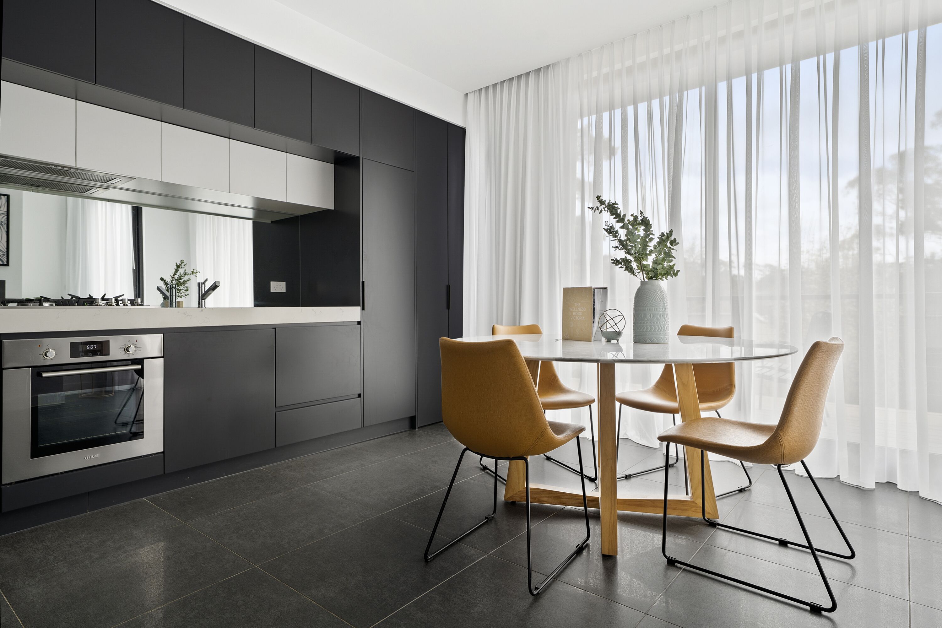 A modern kitchen with a built-in oven, a dining table with four chairs, and a large window with sheer curtains.