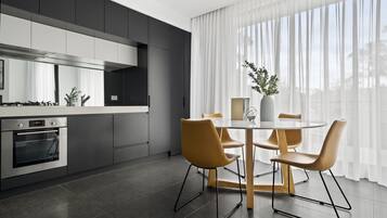 A modern kitchen with a built-in oven, a dining table with four chairs, and a large window with sheer curtains.