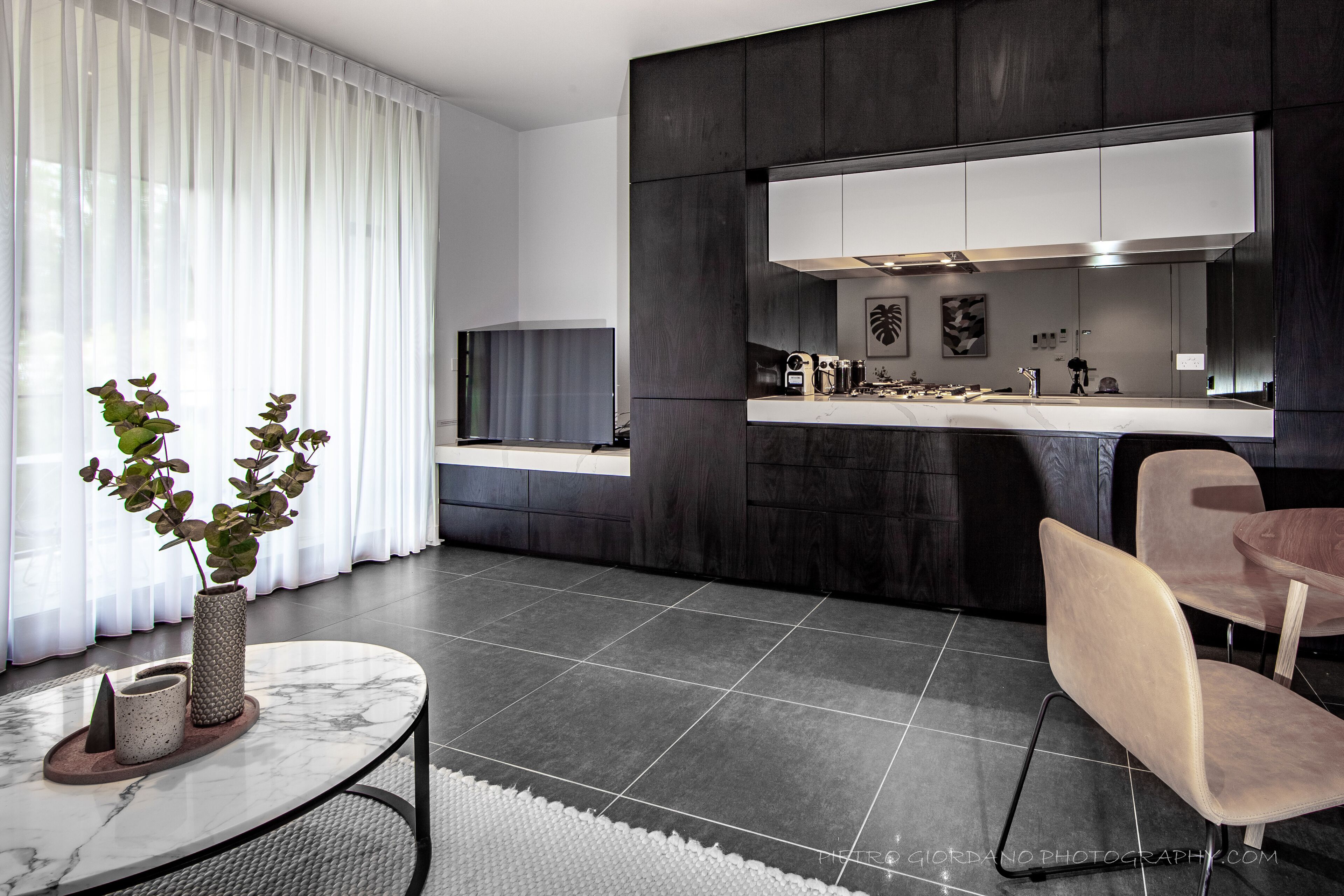 A modern kitchen with dark cabinetry, a white countertop, and a large window with sheer curtains.