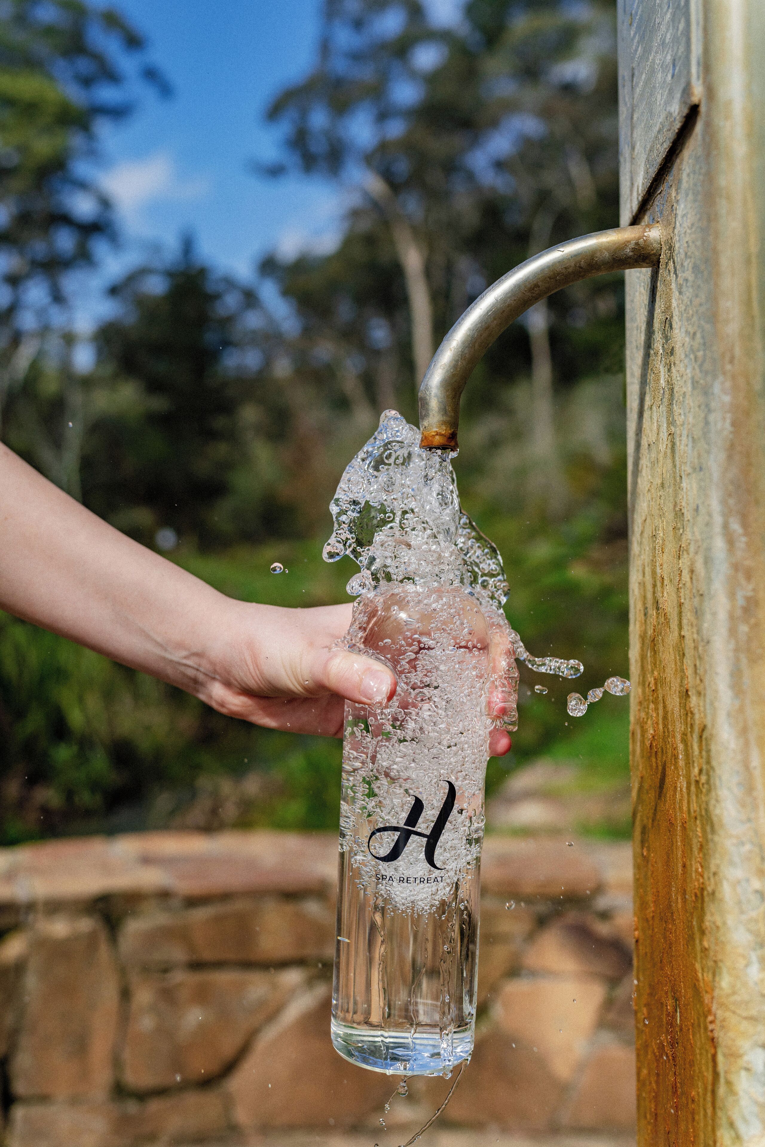 Une main tenant une bouteille en verre d’où s’écoule de l’eau par un bec verseur en métal.