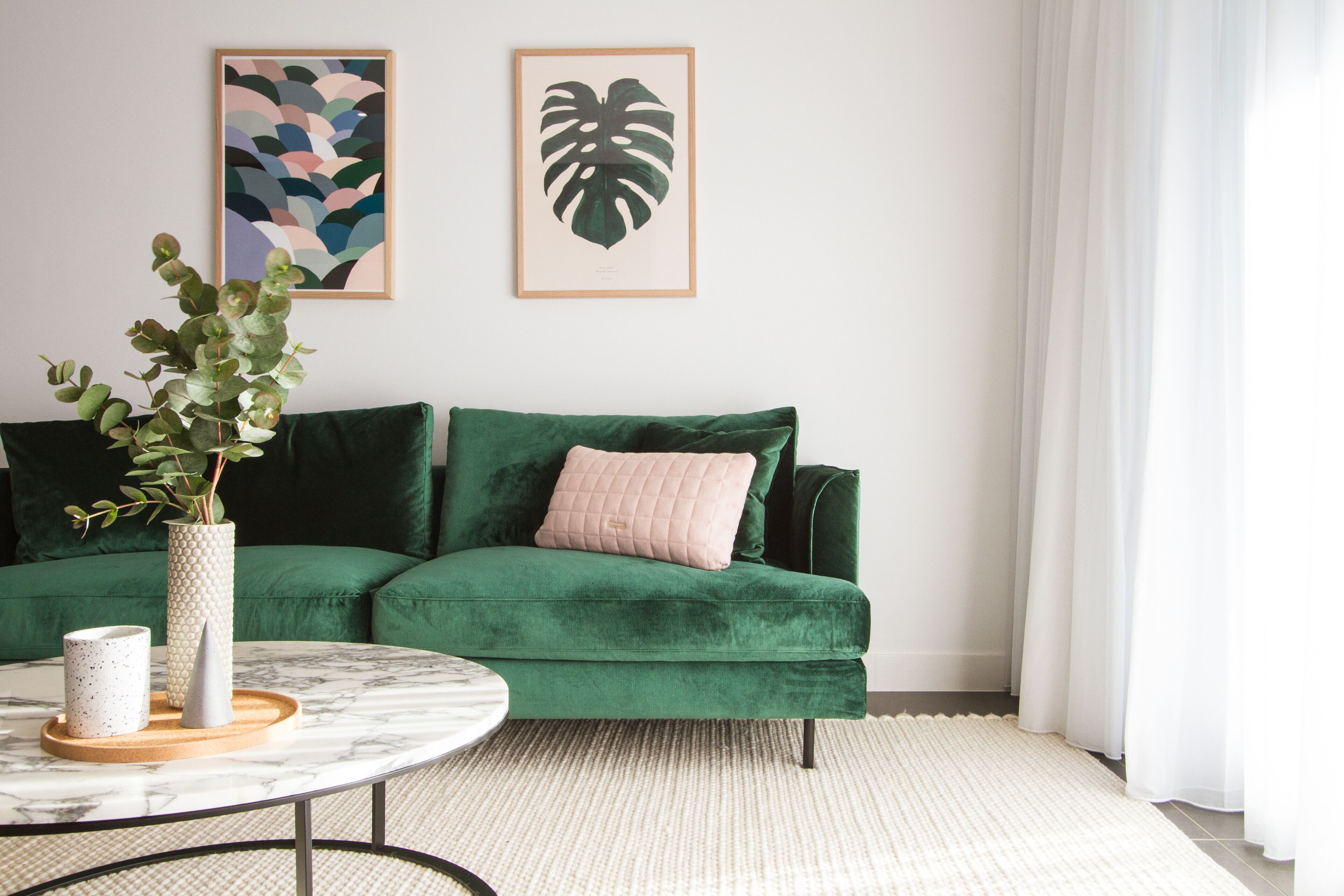 A green sofa with a pink pillow, a white vase with green leaves, and a marble coffee table with a tray and a white mug.