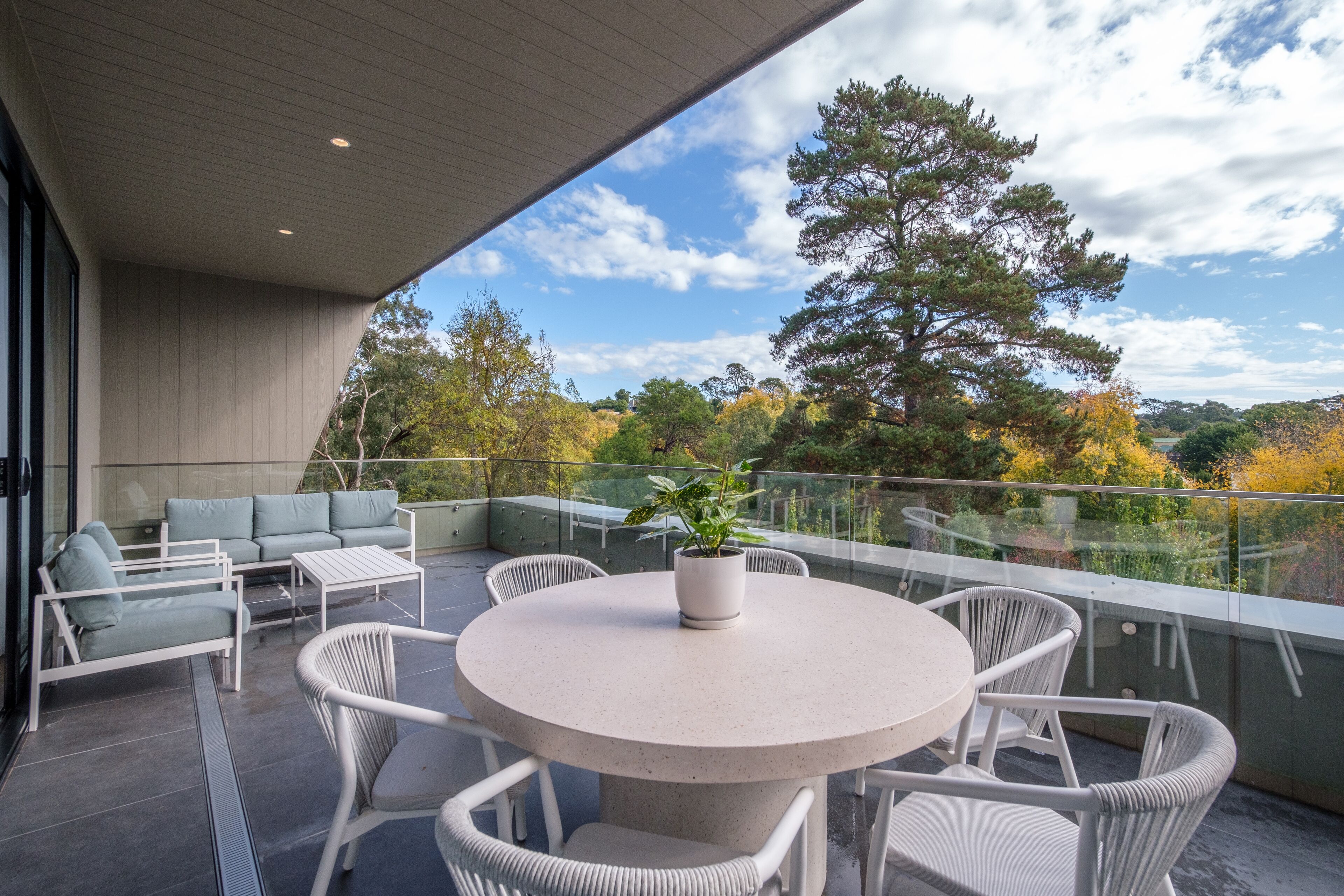 A modern outdoor patio with a round table and white wicker chairs, overlooking a scenic view of trees and hills.