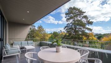 A modern outdoor patio with a round table and white wicker chairs, overlooking a scenic view of trees and hills.