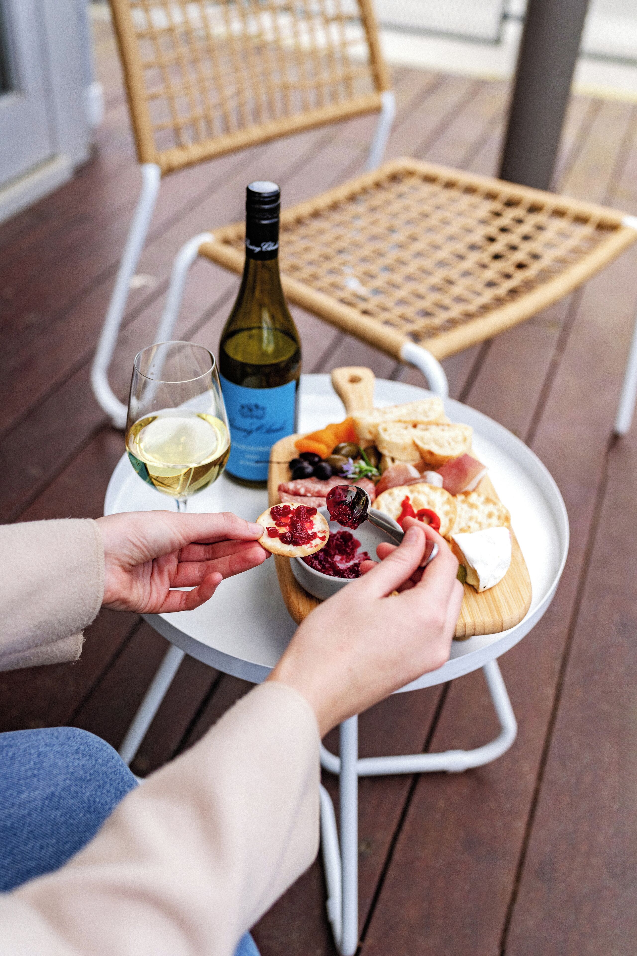 Una persona sosteniendo un trozo de queso y una copa de vino blanco sobre una mesa redonda, con una botella de vino, un plato de queso, pan y aceitunas.