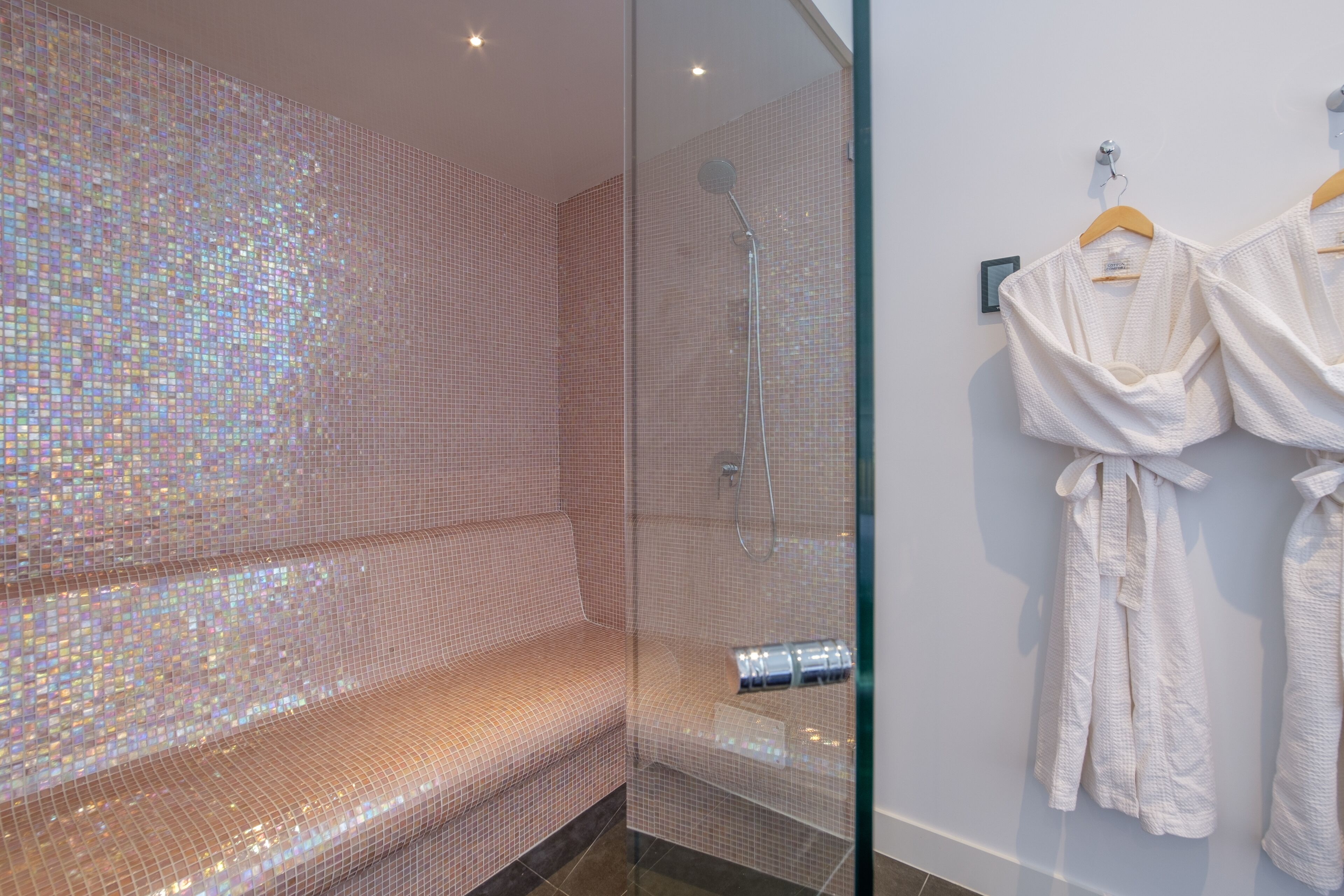 A modern bathroom with a glass-enclosed shower, a seating area with a mosaic tile wall, and two white robes hanging on the wall.