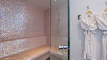 A modern bathroom with a glass-enclosed shower, a seating area with a mosaic tile wall, and two white robes hanging on the wall.