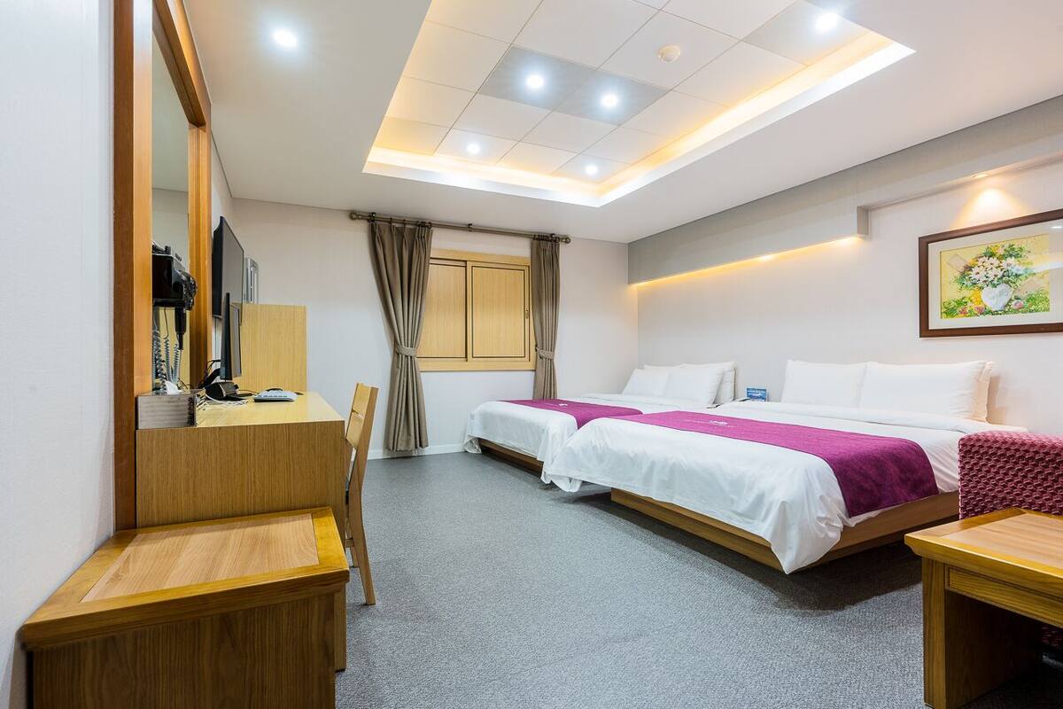 A hotel room with a desk, two beds, a chair, a window with curtains, and a ceiling light.