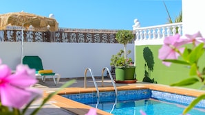 A swimming pool with a tiled edge, a green chair, a thatched umbrella, and a potted plant with flowers. - Hearth away from house BREIA (Tías)