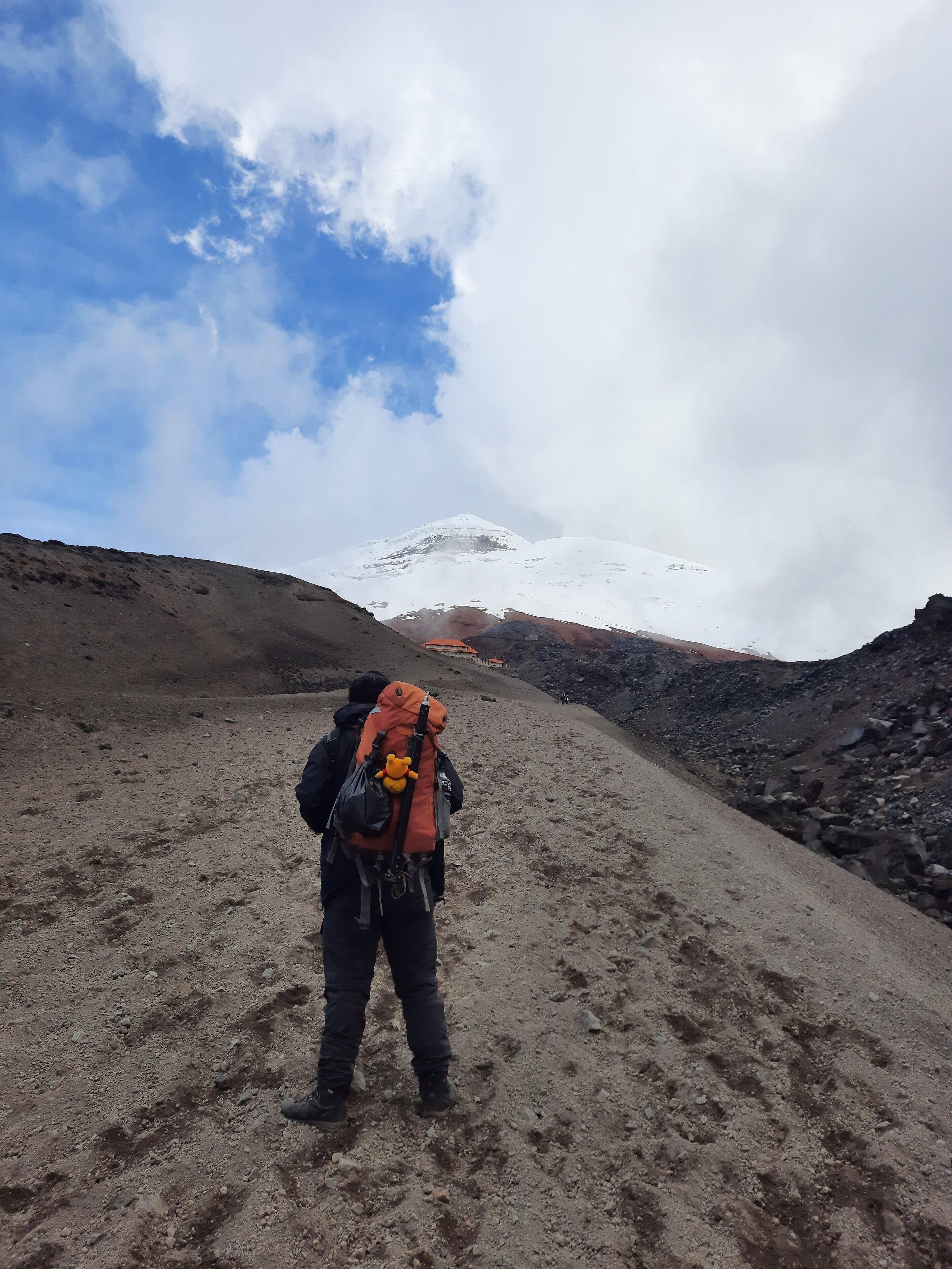 Un excursionista con una mochila y un peluche en la espalda, caminando por un sendero rocoso hacia una montaña nevada.