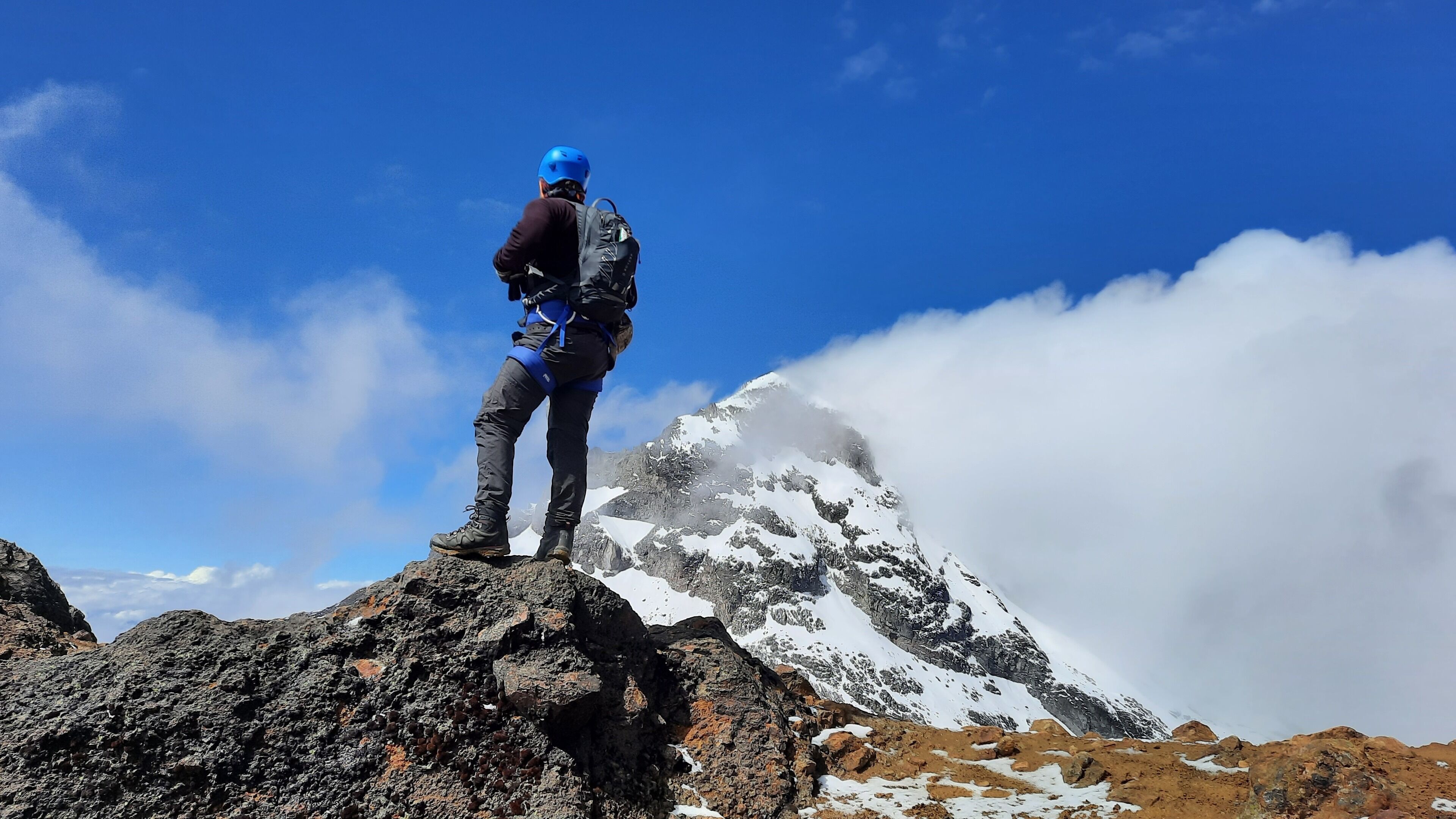 Una persona de pie sobre una cima rocosa, con una montaña al fondo.