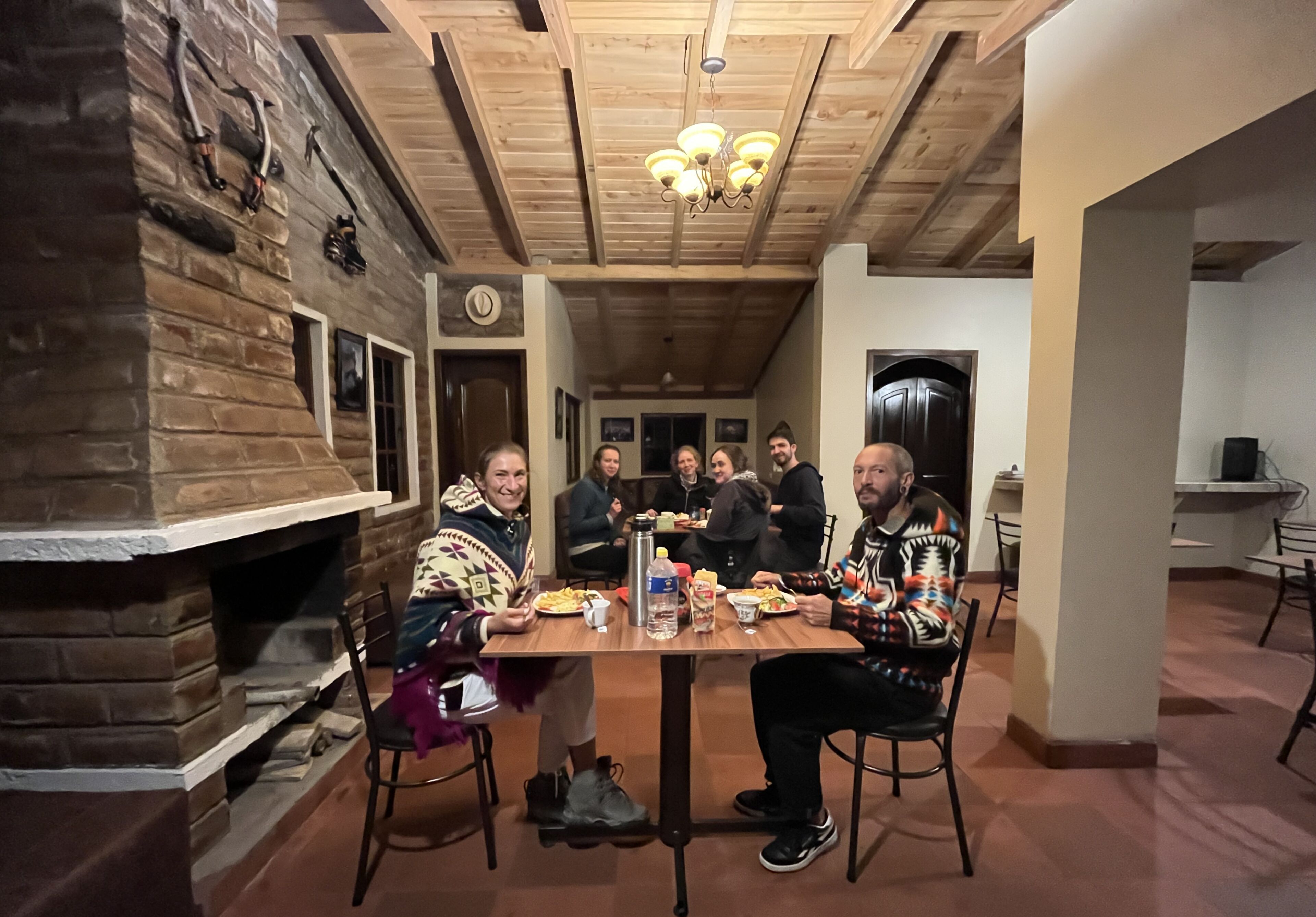 Eine Gruppe von Personen speist an einem Holztisch in einem Raum mit Kamin aus Stein und Holzbalkendecke.