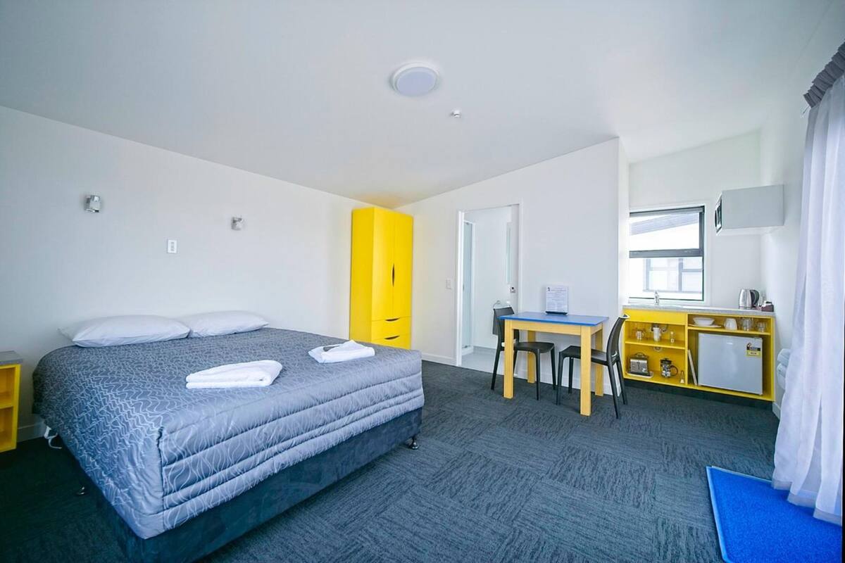 A hotel room with a bed, a desk, a chair, a small kitchenette, and a bathroom.