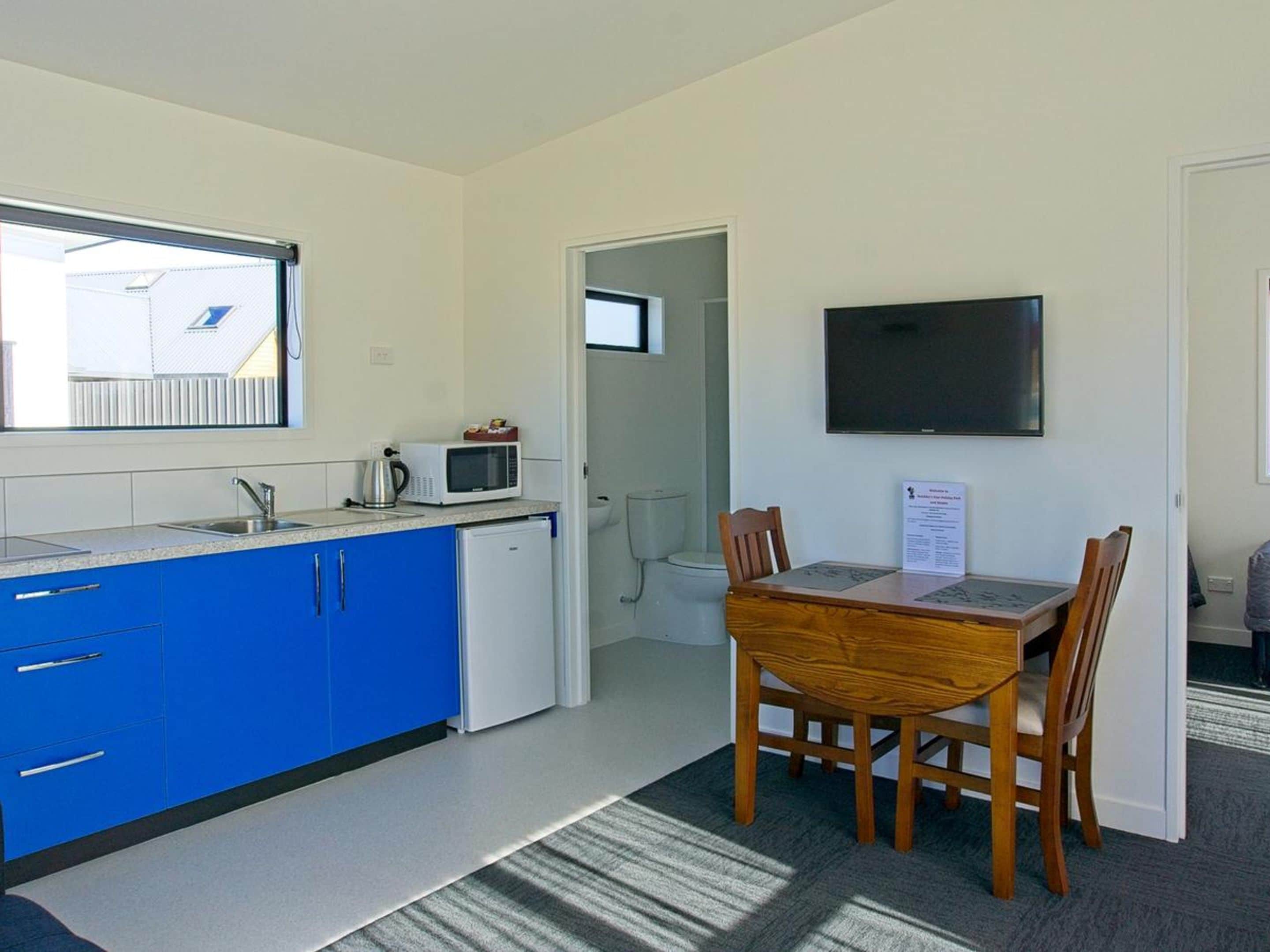 Una cocina compacta con armarios azules, un refrigerador blanco, un microondas y una tetera. Hay una mesa de comedor con cuatro sillas y un televisor de pantalla plana en la pared.