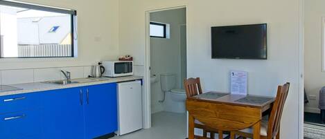Una cocina compacta con armarios azules, un refrigerador blanco, un microondas y una tetera. Hay una mesa de comedor con cuatro sillas y un televisor de pantalla plana en la pared.