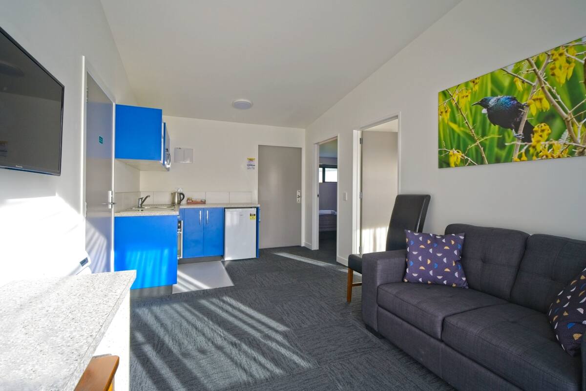 A modern living room with a grey sofa, a coffee table, and a painting of a bird. There is a kitchen area with blue cabinets and a refrigerator.