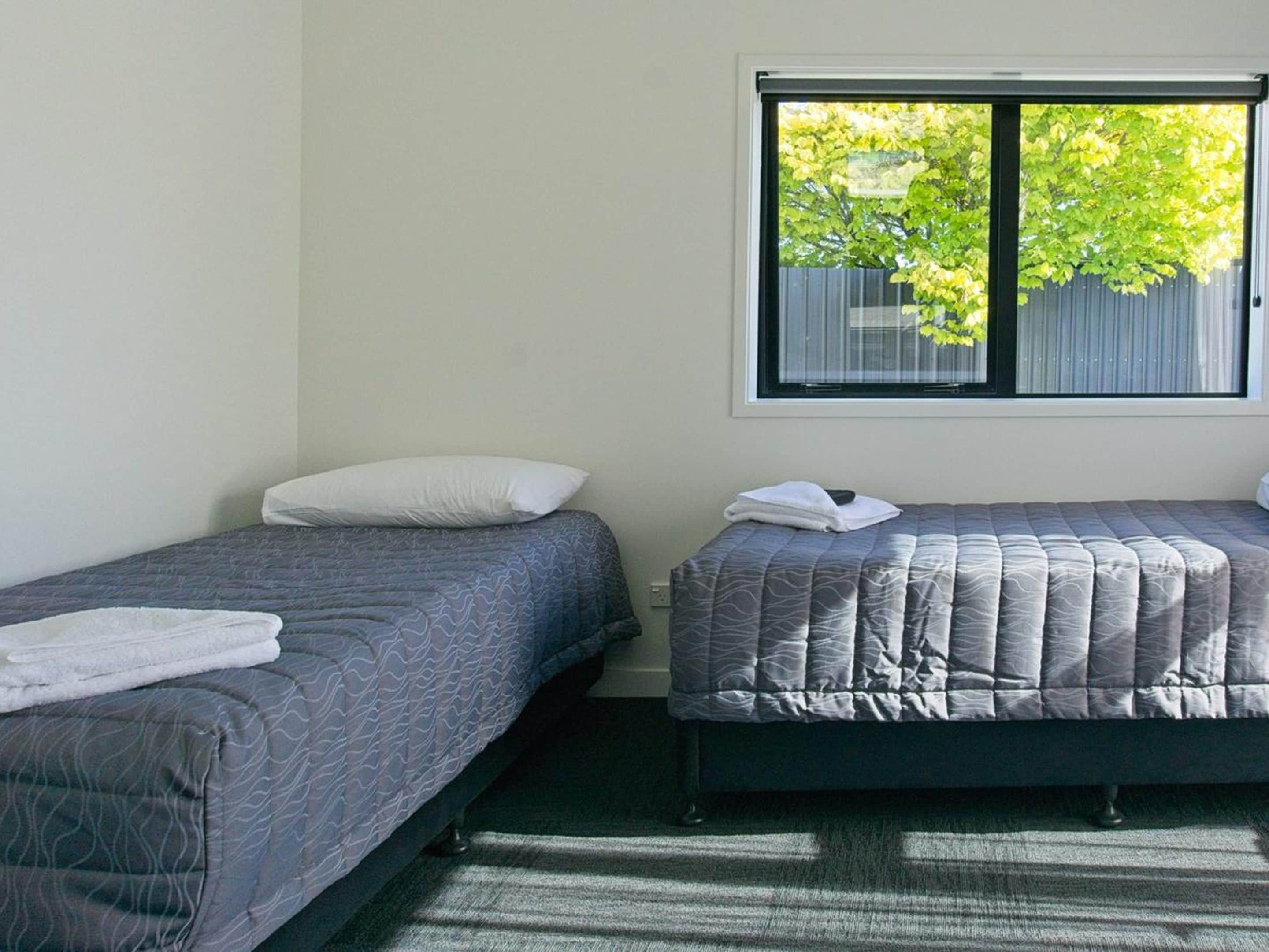 Une chambre avec deux lits simples, chacun recouvert d’une couette à motifs gris et agrémenté d’oreillers blancs. Une fenêtre offre une vue sur les arbres et une clôture.