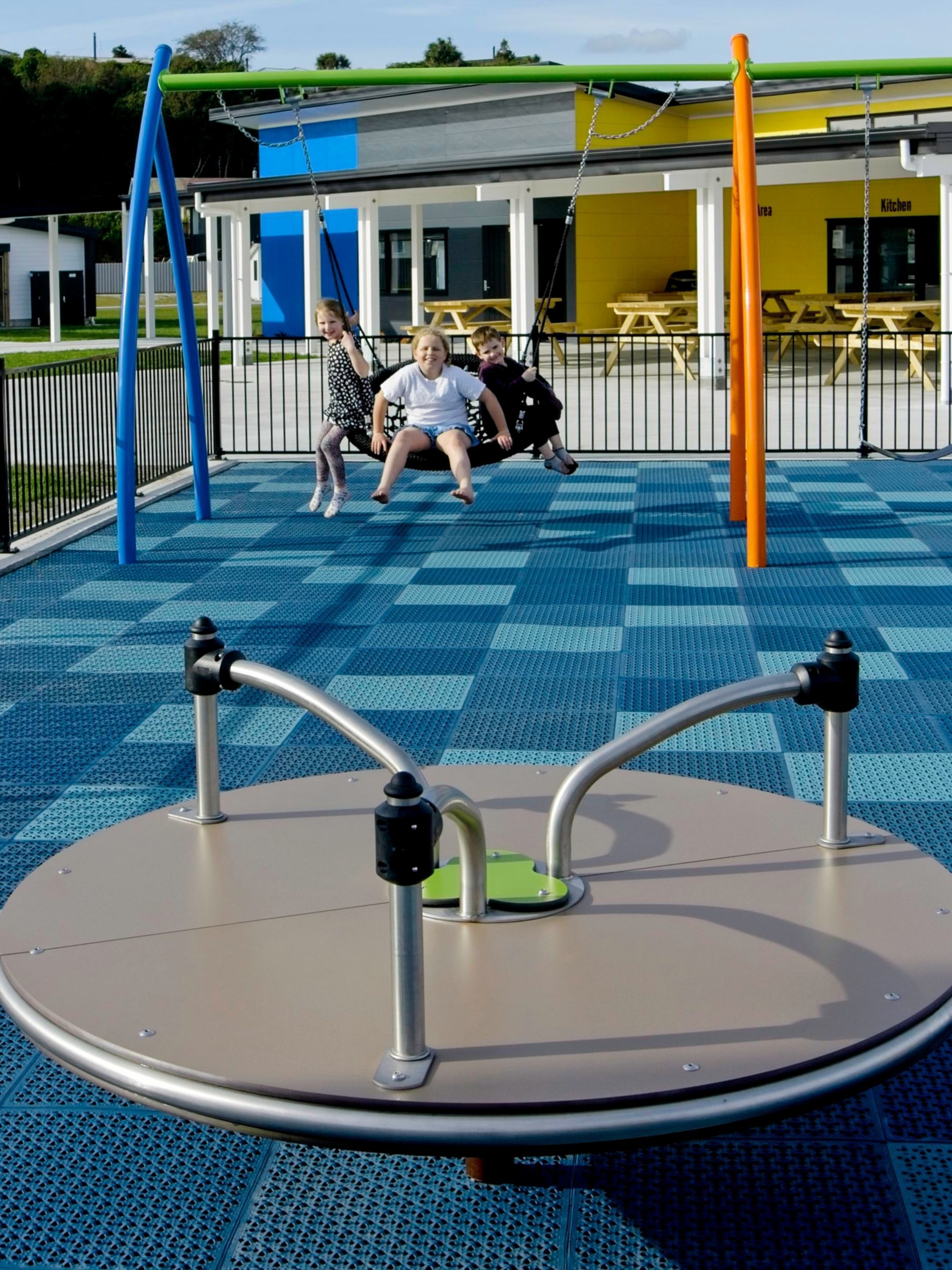 A playground with a blue checkered floor, a circular play structure, and a building with a yellow exterior in the background.