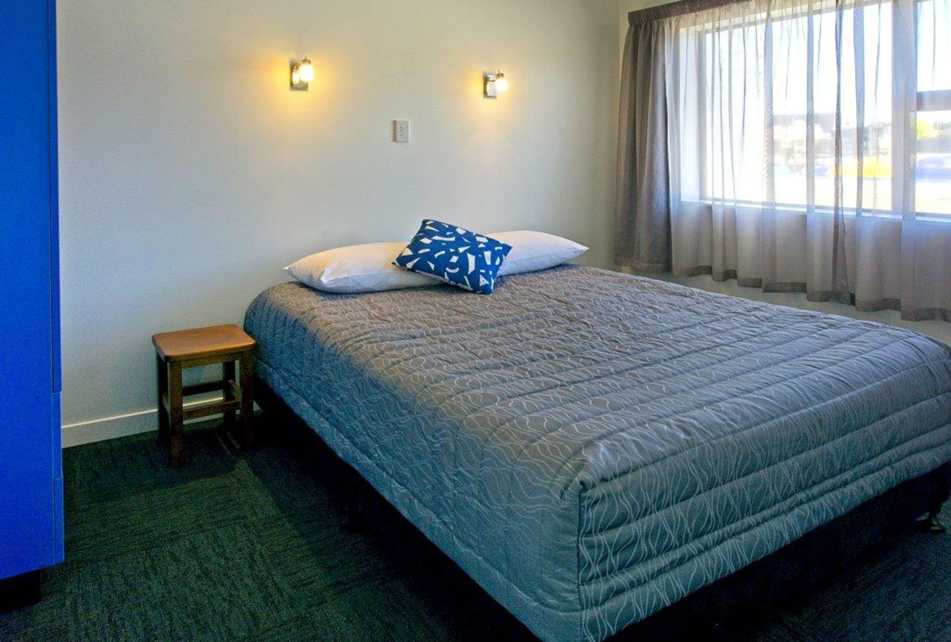 A single bed with a patterned bedspread, two wall-mounted lights, a small wooden stool, and a window with sheer curtains.