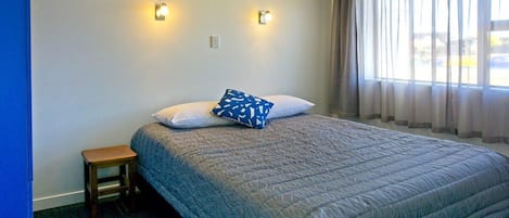Una cama individual con una colcha estampada, dos lámparas de pared, un taburete pequeño de madera y una ventana con cortinas traslúcidas.
