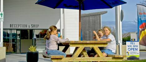 Ein Holzbistro-Tisch mit einem blauen Sonnenschirm vor einem Gebäude, auf dem 'RECEPTION & SHOP' steht.