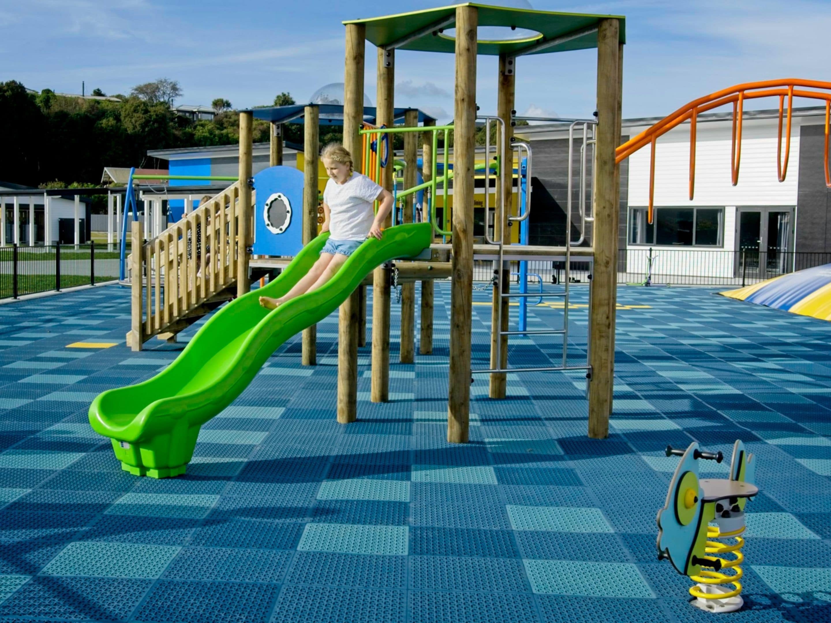 Un parque infantil con un tobogán verde, estructuras para escalar y un suelo azul a cuadros.