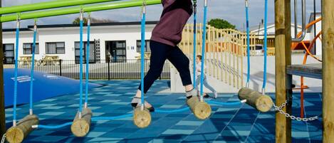 Un enfant joue dans un parc, sur une aire de jeux dotée d’un sol à carreaux bleus et d’une structure d’escalade verte.