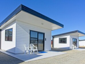 Un motel moderno de una sola planta con revestimiento blanco y techo oscuro. Hay dos sillas de plástico blanco fuera de la entrada.