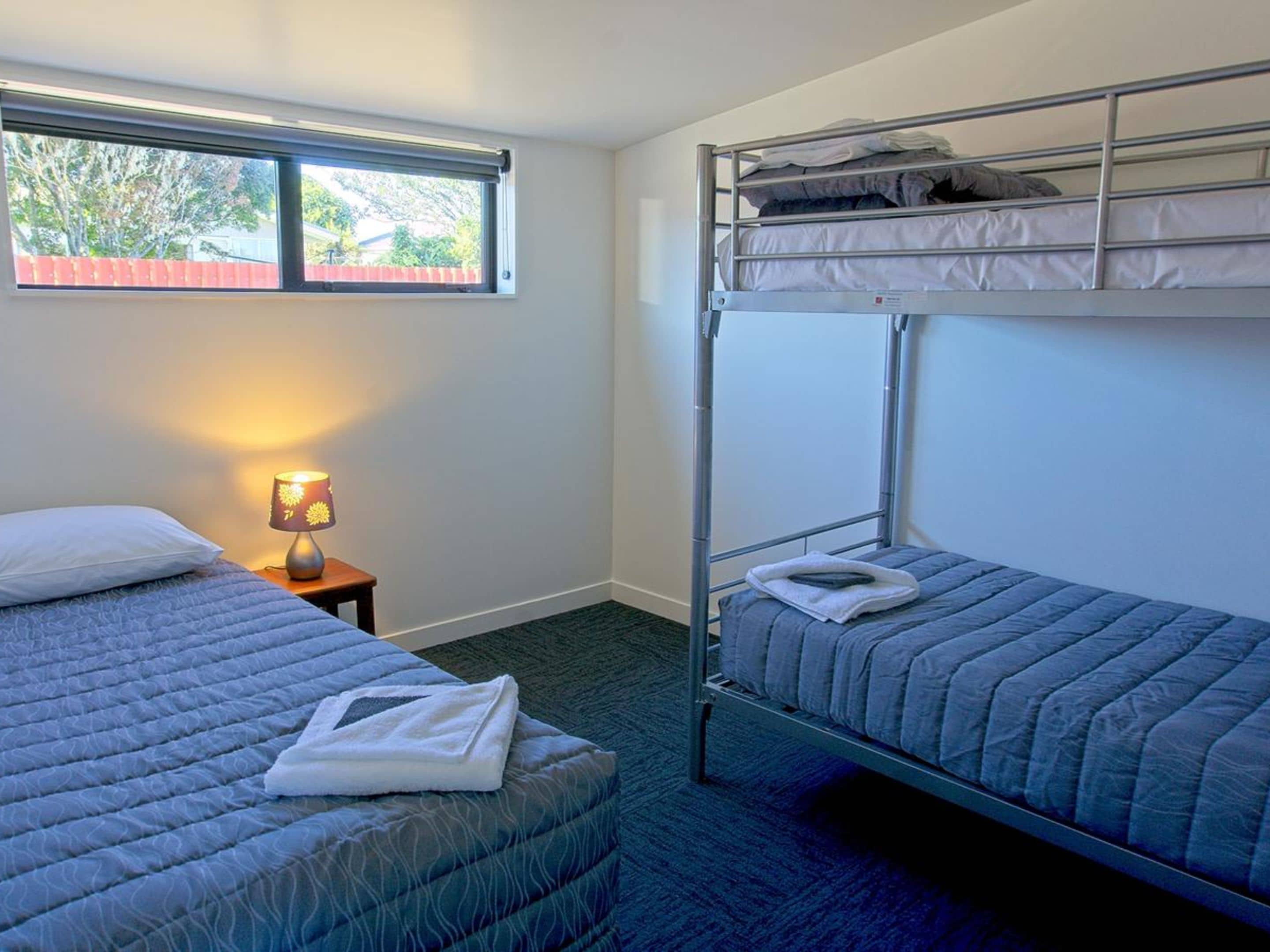 Habitación de hotel con literas, una cama individual, una mesita de noche con lámpara y ventana con vista a árboles.