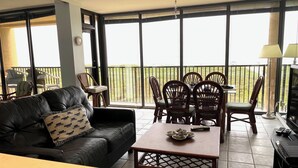 A living room with a black leather sofa, a wooden dining table with chairs, and a balcony with a view.