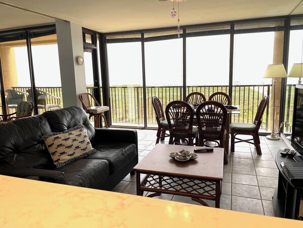 A living room with a black leather sofa, a wooden dining table with chairs, and a balcony with a view.