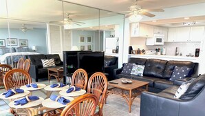 A spacious living area with a dining table set for four, surrounded by wicker chairs. A dark leather sofa and a flat-screen TV are visible in the background.