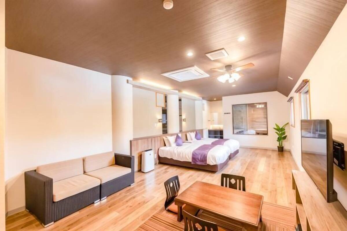 A modern hotel room with a bed, sofa, dining table, and a ceiling fan.