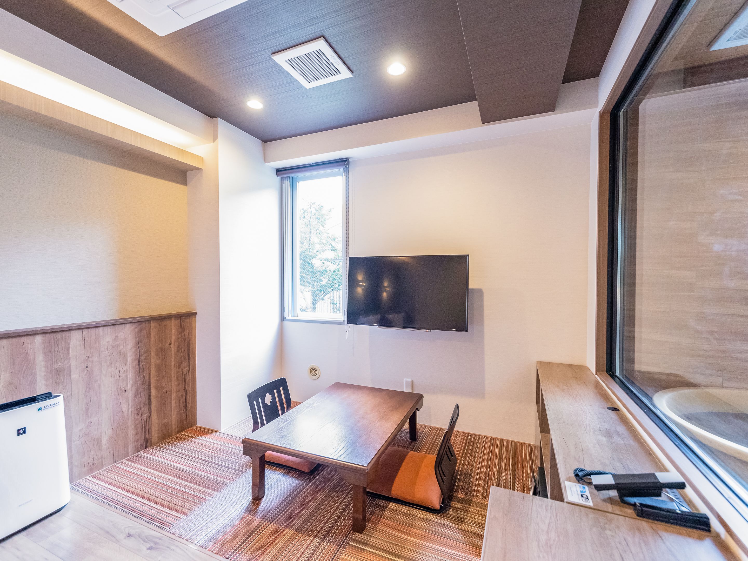 Una habitación moderna con una mesa de madera, sillas, un televisor de pantalla plana en la pared y una ventana que proporciona luz natural.