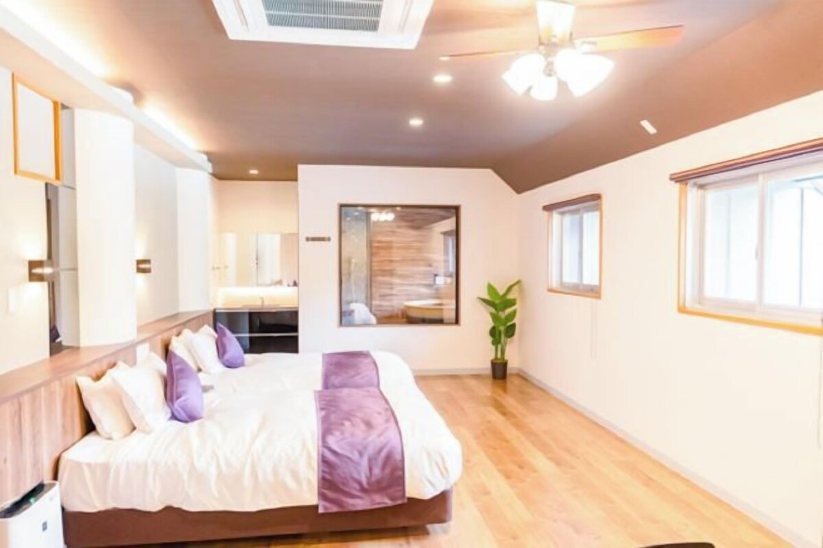 A modern hotel room with a large bed, a ceiling fan, a potted plant, and a large window.
