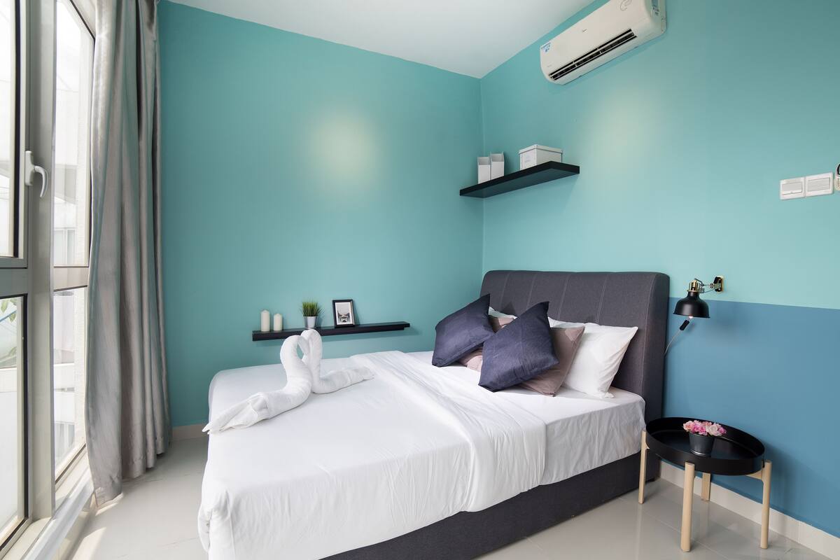 A modern bedroom with teal walls, a large bed, a bedside table with a lamp, and a window with curtains.