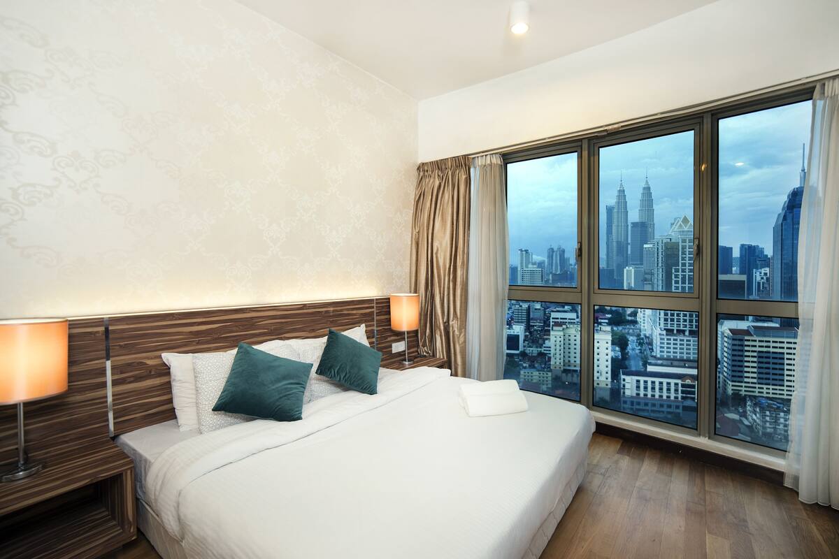 A hotel room with a large bed, two bedside tables with lamps, a view of a cityscape through large windows, and a patterned wall.