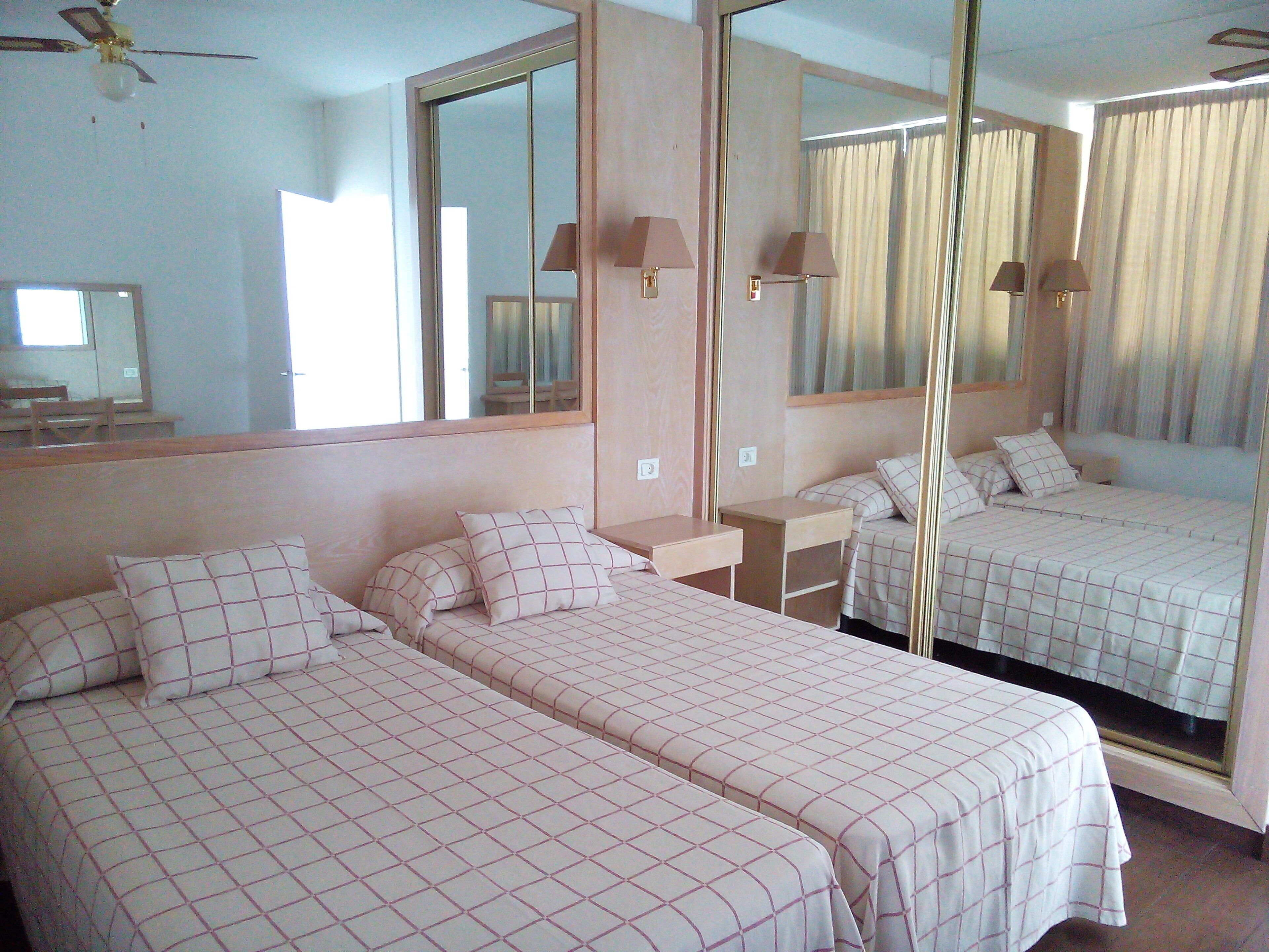 A hotel room with two single beds, a large mirror, a ceiling fan, and a window with curtains.