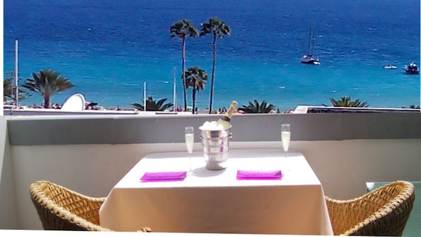 Una mesa con una botella de champán, dos copas y un mantel individual morado en un balcón con vista al océano, barcos y palmeras.